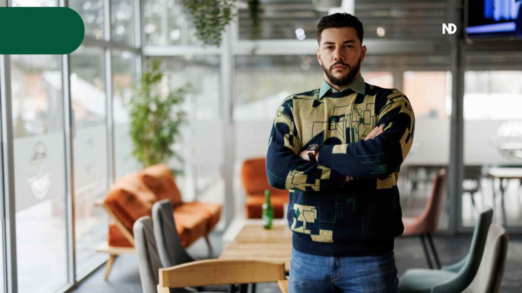 Hombre joven de pie saco con diseño, camisa y jeans, barba, fondo muebles grises y naranjas planta y ventanal grande 