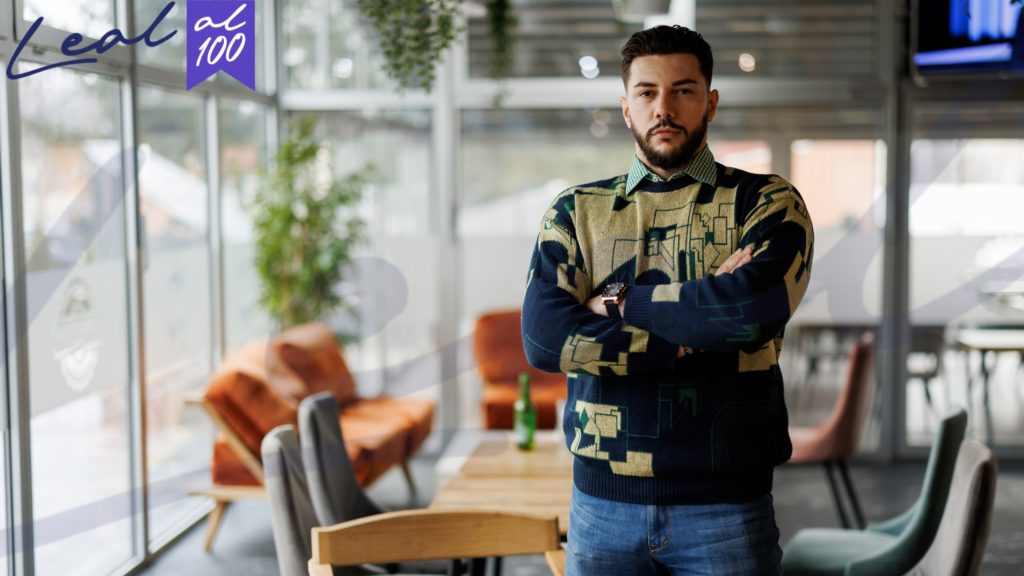 Hombre joven de pie saco con diseño, camisa y jeans, barba, fondo muebles grises y naranjas planta y ventanal grande