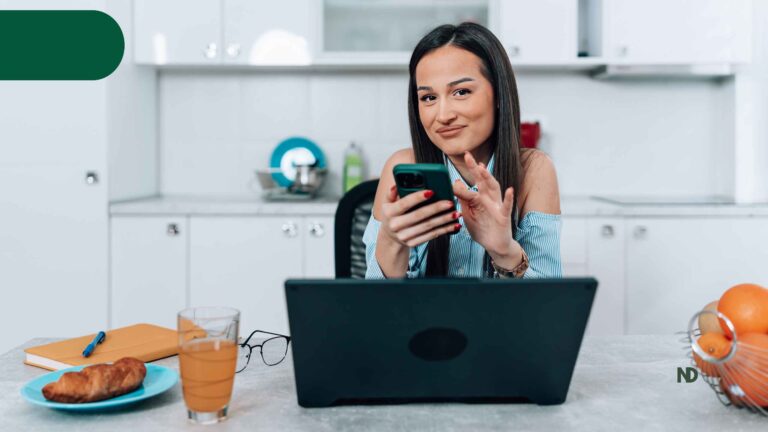 Mujer revisa su teléfono y está sentada frente a su computadora en espacio personal, cocina, jugo y frutas, pan libreta y esfero nómadas digitales españa